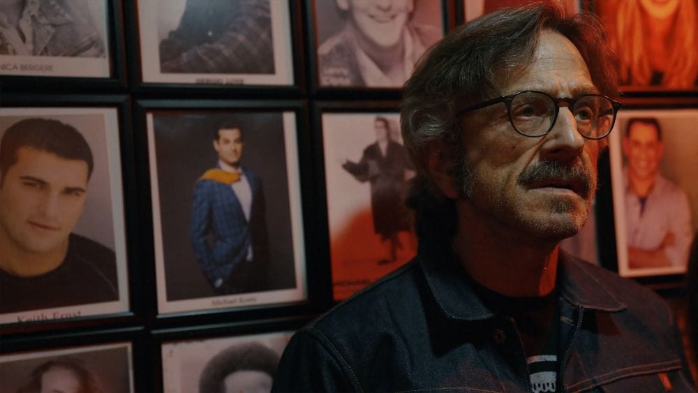 Marc Maron stands in front of a wall of framed comedian photographs inside a comedy club.