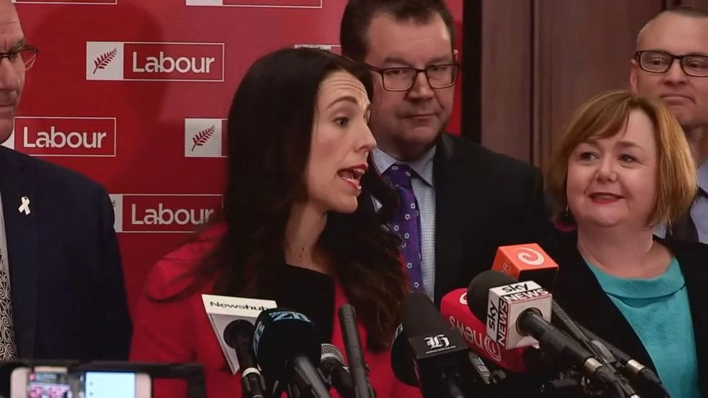 Jacinta Ardern stands in front a red New Zealand Labour Party sign surrounded by other politicians, microphones in her face.