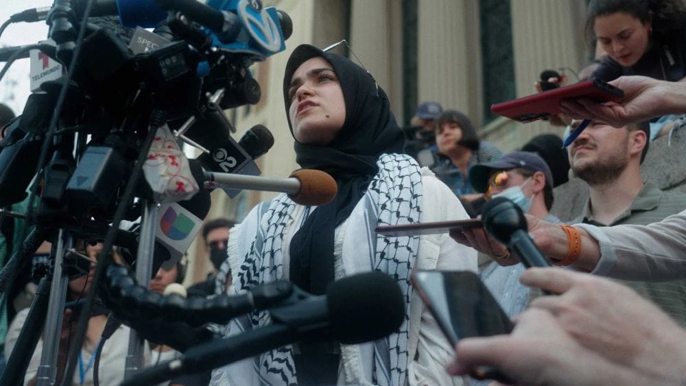 A young woman wearing a Palestinian keffiyeh and head-scarf stands on the steps of an old building in front of microphones.