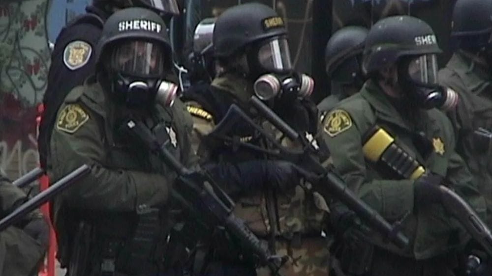 police and sheriffs in riot gear and gas masks holding guns to shoot tear gas at protesters.