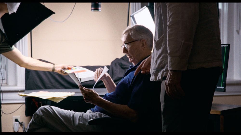 A man with gray hair and glasses looks at paperwork with camera and lighting around him.