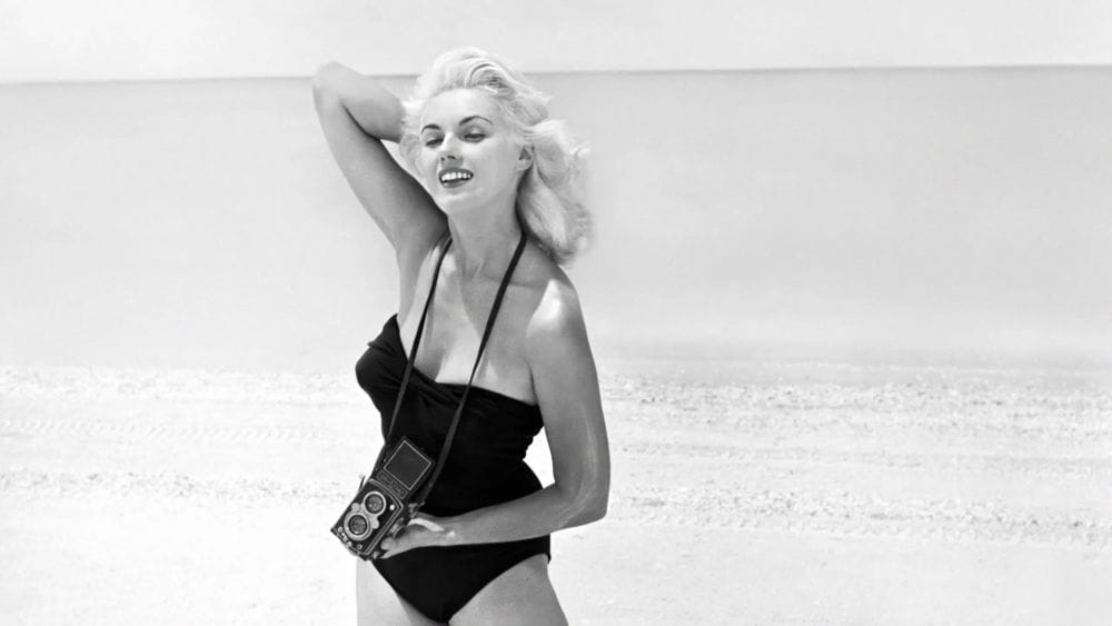 A blonde woman in a black swimsuit stands on a beach holding a camera.