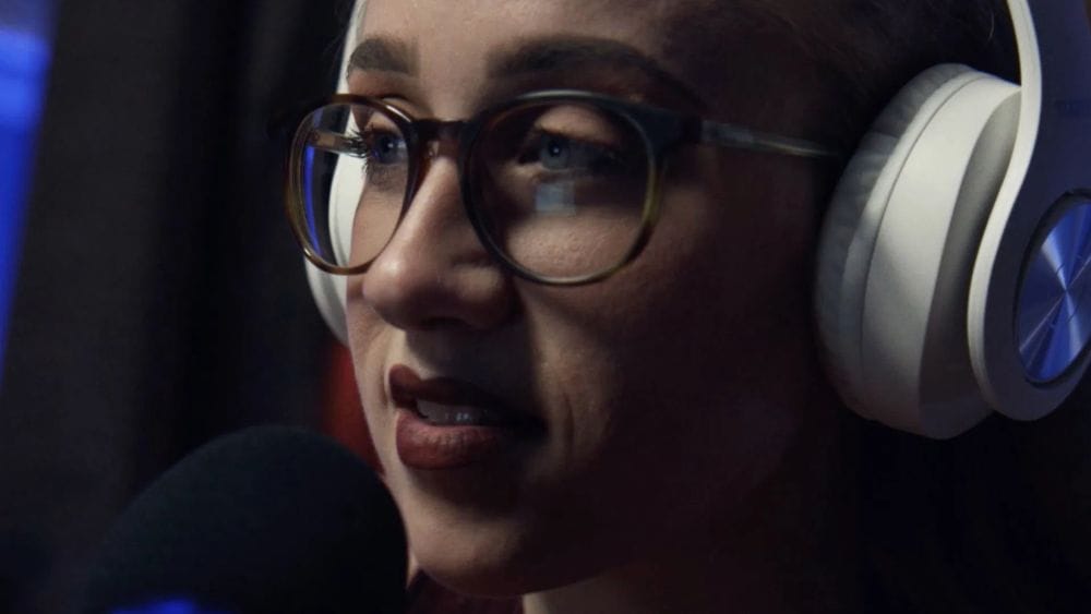 A young woman with glasses and large white headphones speaks into a microphone at night.