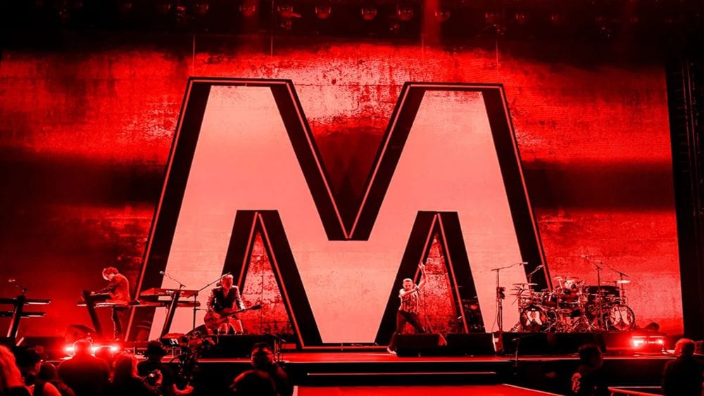 A concert stage bathed in red lighting featuring a large letter M and a band in front it.