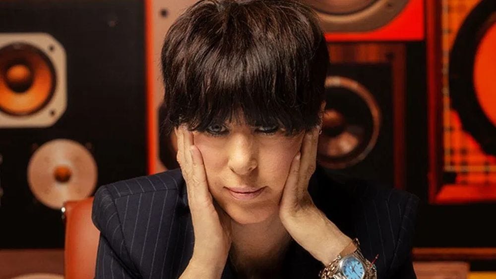 Diane Warren sits in front of a wall of speakers and stares directly into the camera with her hands on her face.