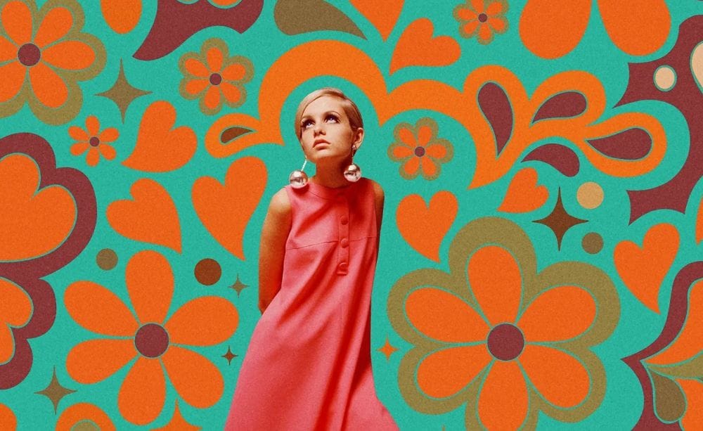 A slim woman with short blonde hair, big bauble earrings and a pink dress stands amid a stylised 1960s flower pattern.