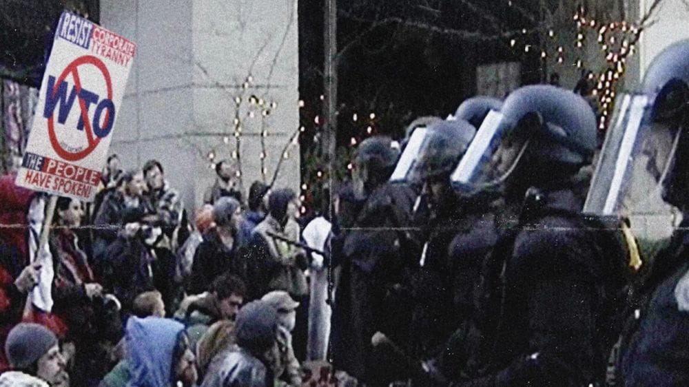 Protesters hold a sign that reads resist corporate tyranny the people have spoken and WTO with a red line through it in front of police officers in masks and riot gear.