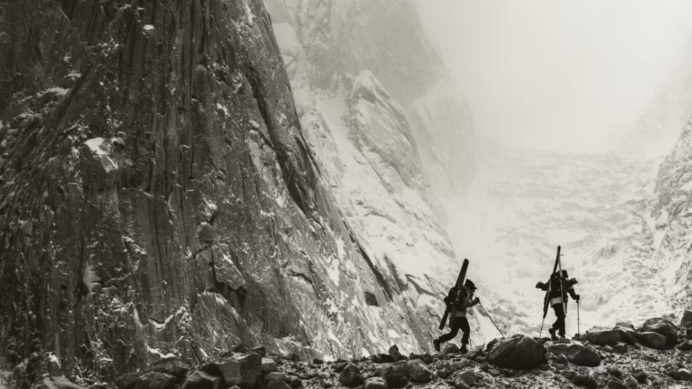 Two people hike with skis on their face as massive mountain faces appear behind them.