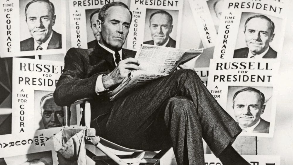 Henry Fonda in a suit and tie sits cross-legged reading a newspaper surrounded by signs that read Russell for President, a time for courage.