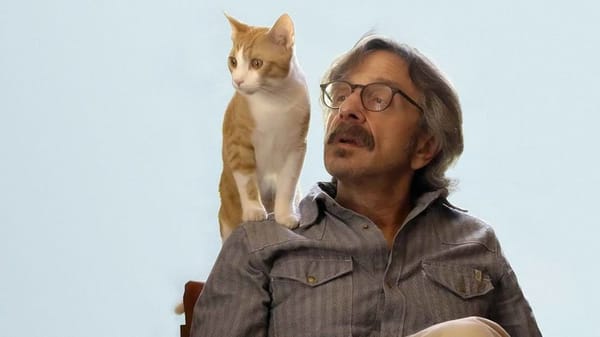 Marc Maron with glasses and a thick moustache sits while a ginger and white cat sits on his shoulder.