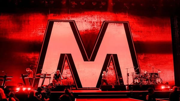 A concert stage bathed in red lighting featuring a large letter M and a band in front it.