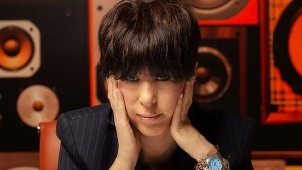 Diane Warren sits in front of a wall of speakers and stares directly into the camera with her hands on her face.