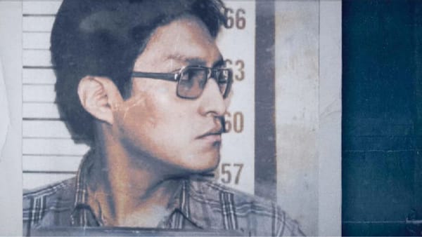 An Apache man in glasses and a plaid shirt stands in front of a police height chart looking to his side.