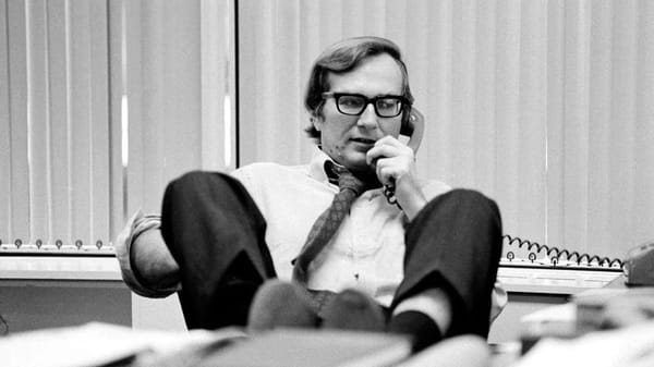 Black and white photo of a man in white business shirt, tie and dark slacks, leaning back in a chair while on the telephone.
