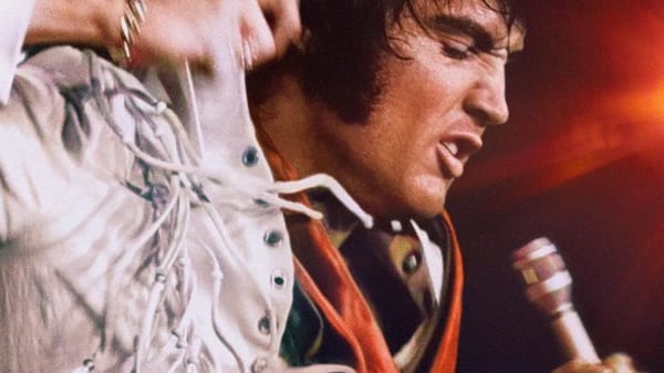 Elvis Presley in a white jumpsuit sings into a microphone, an intense look on his face.