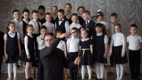 A teacher in black adjusts a microphone in front of a classroom choir of young children.