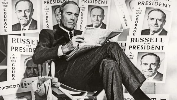 Henry Fonda in a suit and tie sits cross-legged reading a newspaper surrounded by signs that read Russell for President, a time for courage.